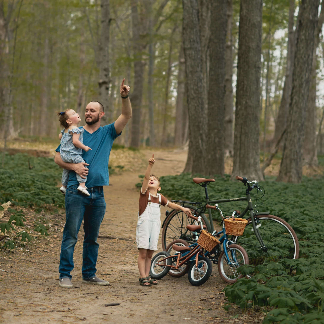 A parent and two children explore a forest path, with bicycles nearby. One child points upward, capturing a moment of wonder.