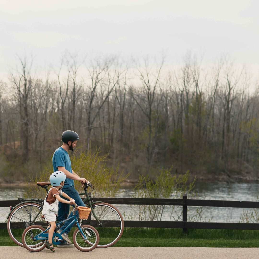 An adult and child ride bikes along a trail with a fence and body of water in the background.