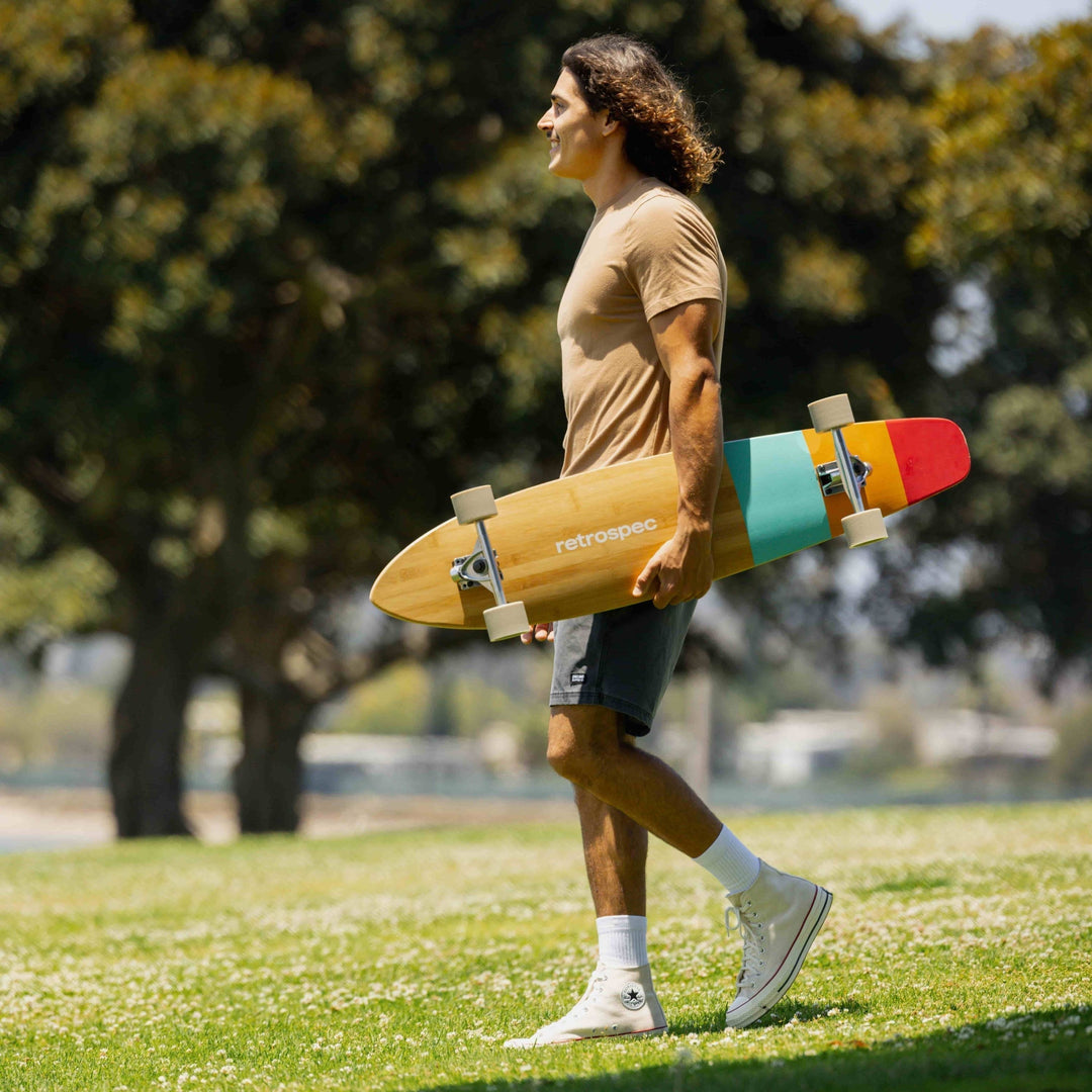A person walks through a grassy park holding a colorful skateboard, dressed in a beige shirt, shorts, and white sneakers.