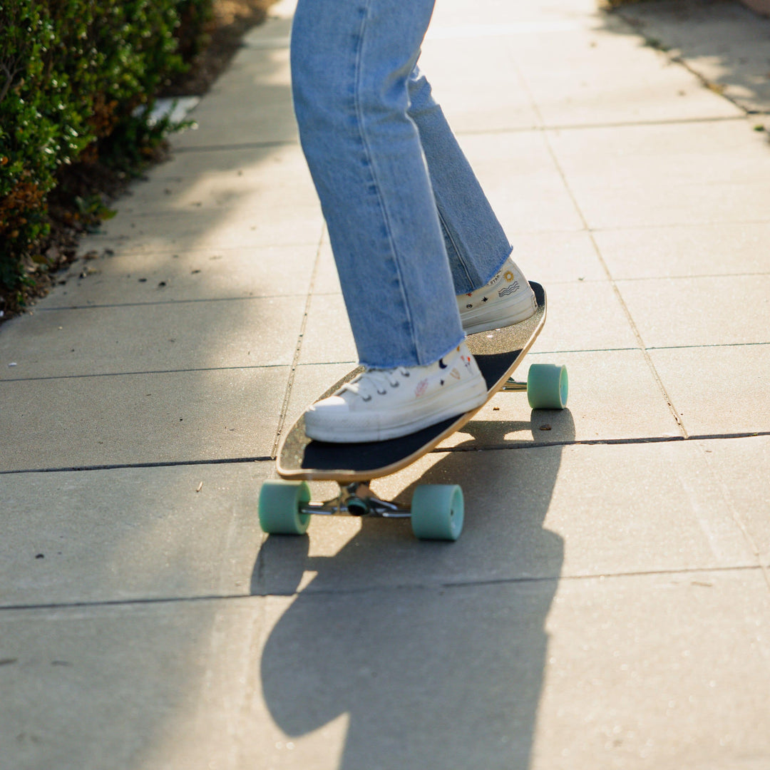 A person wearing jeans and white sneakers rides a skate board with blue wheels down a narrow sidewalk.