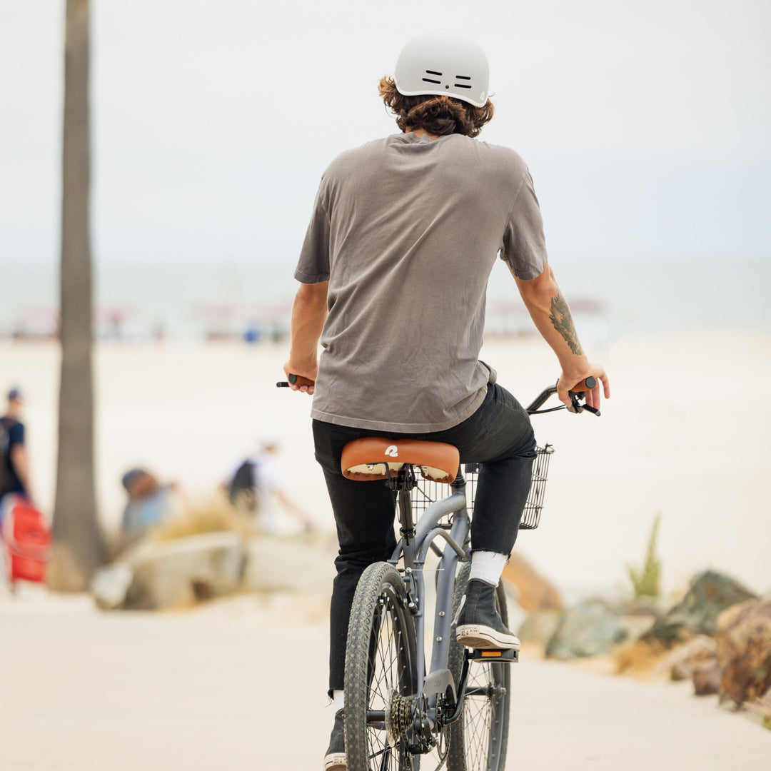 A person in a gray shirt and white helmet rides a bike along a beach path, with ocean and people in the background.