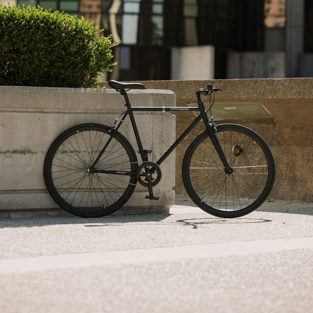 A sleek black retro-style bike rests against a concrete structure, with greenery and modern buildings in the background.