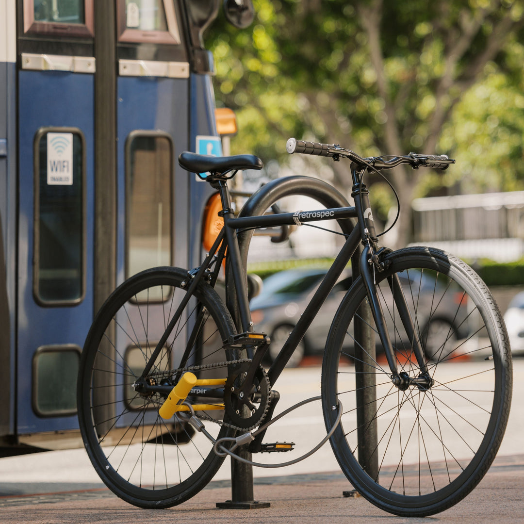 A sleek black bicycle securely locked to a bike rack, with a blue public transit vehicle in the background.