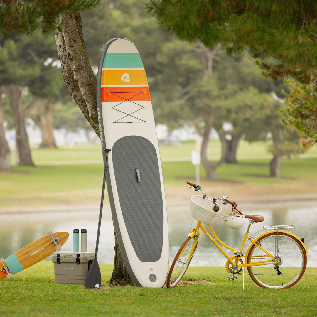 A colorful paddleboard leans against a tree near a yellow bicycle, with a cooler and water bottles on the grass by a serene lake.