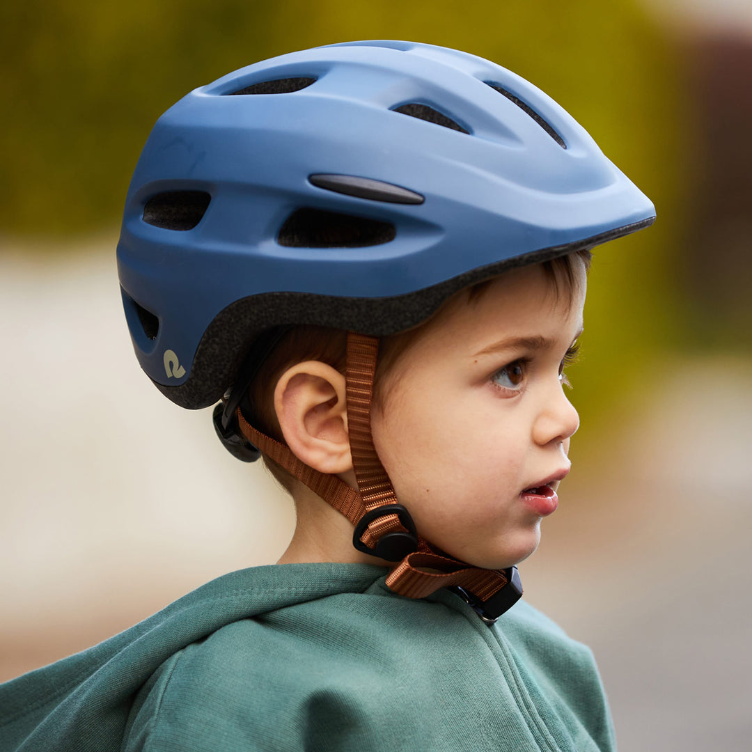 A child wears a blue Scout Kids' Bike & Skate Helmet, secured with a brown strap, while dressed in a green hoodie, outdoors in a blurred background.