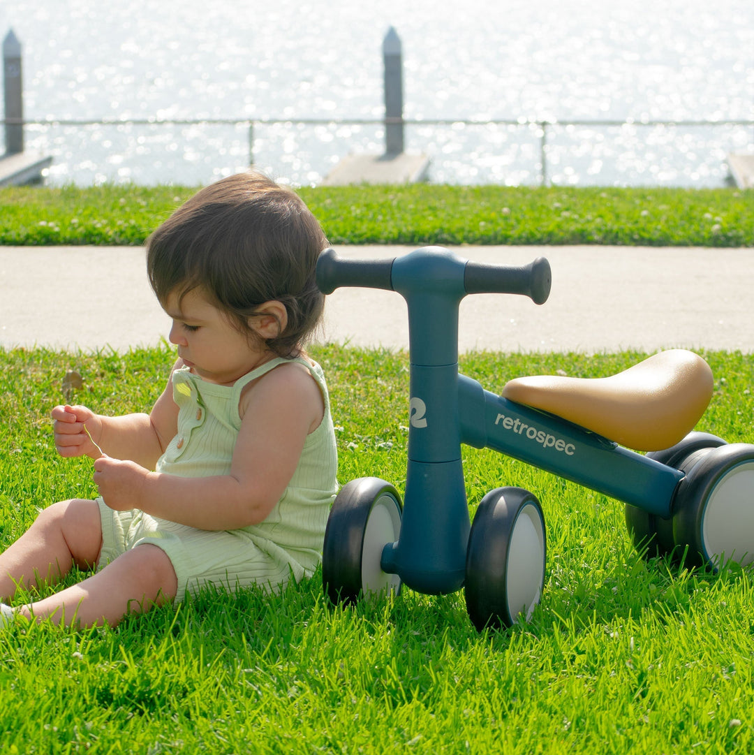 retrospec Cricket 2 teal balance bike with cream seat positioned on grass next to toddler in outdoor setting