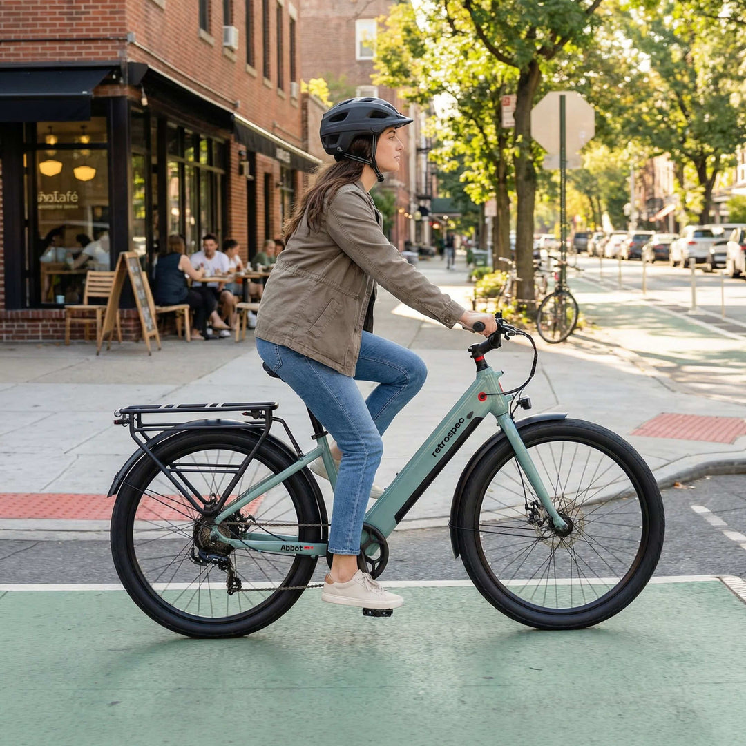 A woman wearing a helmet rides the retrospec Abbot Rev 2 Commuter Electric Bike - Step Through in Sea Glass blue-green through a city bike lane, with tree-lined streets, brick storefronts, and a sidewalk cafe in the background.