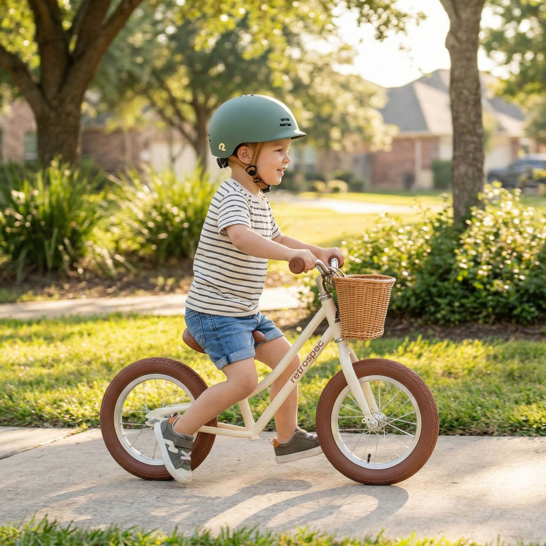 Toddler in a green remi helmet riding a cream retrospec Baby Beaumont toddler balance bike with a wicker basket and brown wall tires on a sunny residential path.
