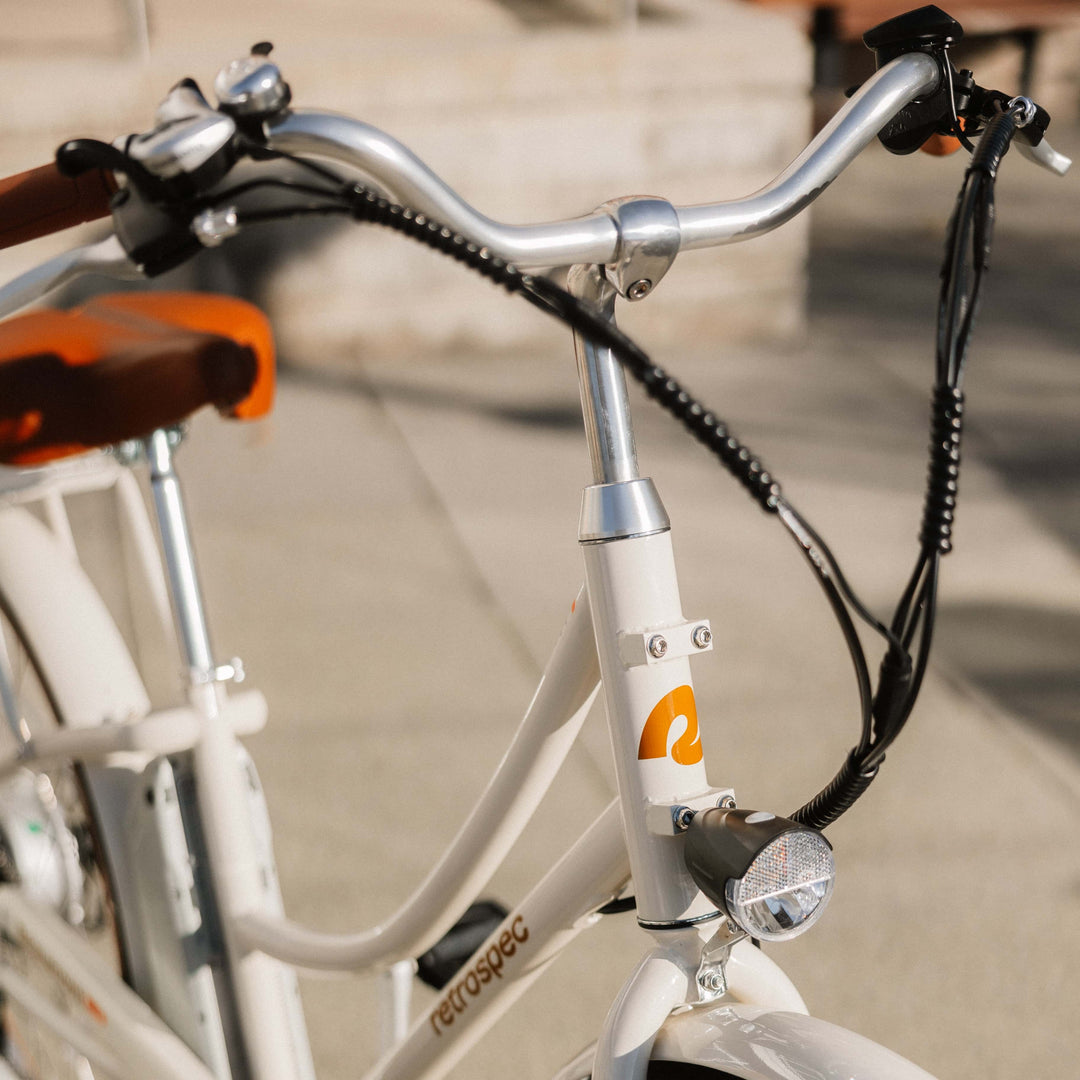 Close-up of a retro-style electric bicycle's handlebars with orange grips, featuring a bell, light, and a white frame with a logo.