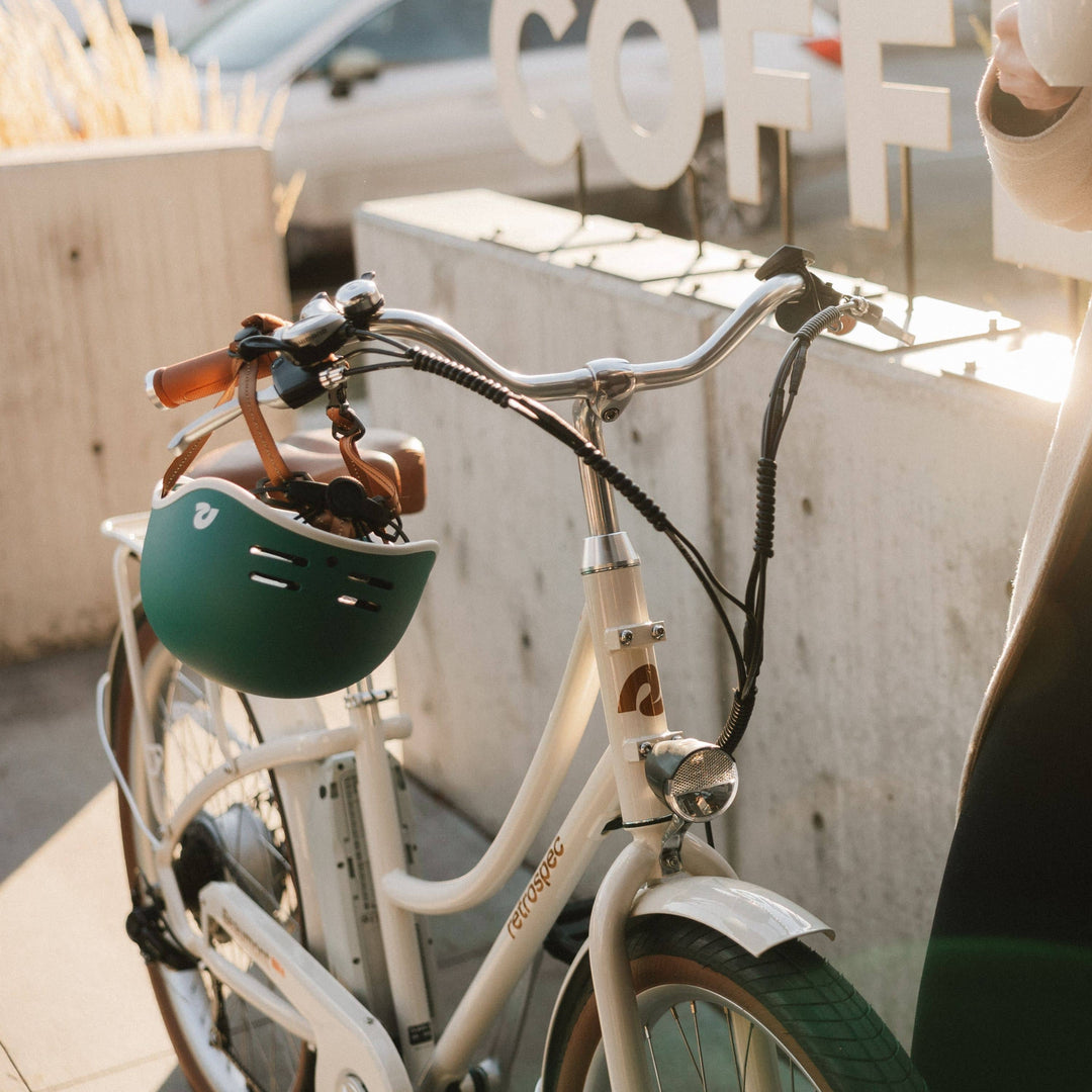 An eggshell Beaumont City Electric Bike rests against a cement outdoor wall with a green helmet hanging by the chinstrap on the bike handle.