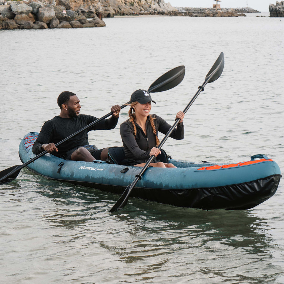 A couple paddle their inflatable tandem kayak on calm water near the shore.
