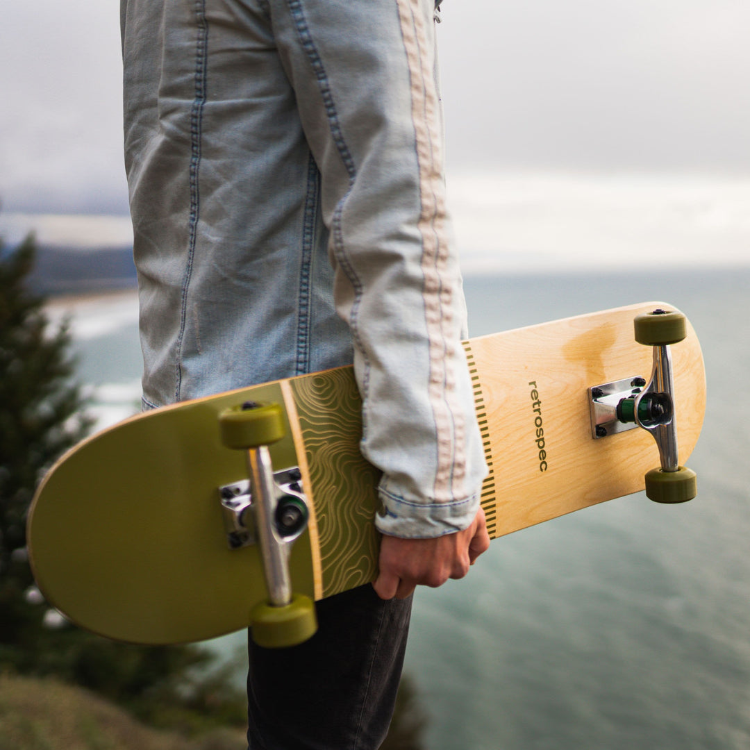 A person wearing a jean shirt holds a skateboard with green wood design with the ocean in the background.