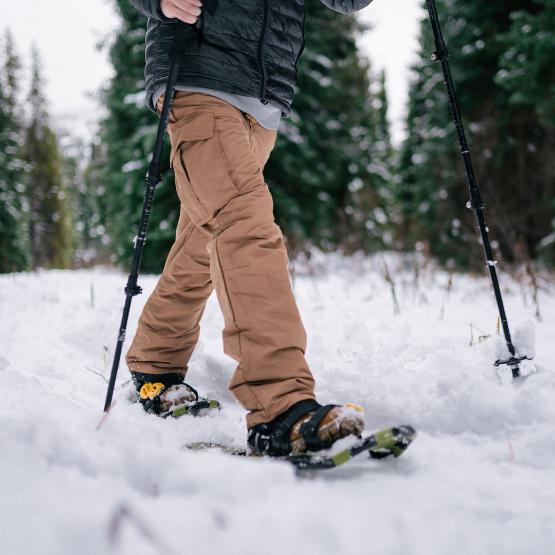 A person snowshoeing through a snowy trail, wearing brown pants and a black jacket, with trekking poles in hand. Pine trees in the background.