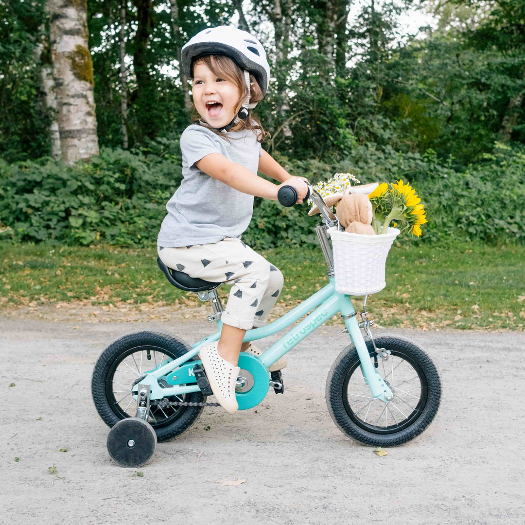 A young child rides a blue bicycle with training wheels and a white basket on the handlebars holds yellow flowers.