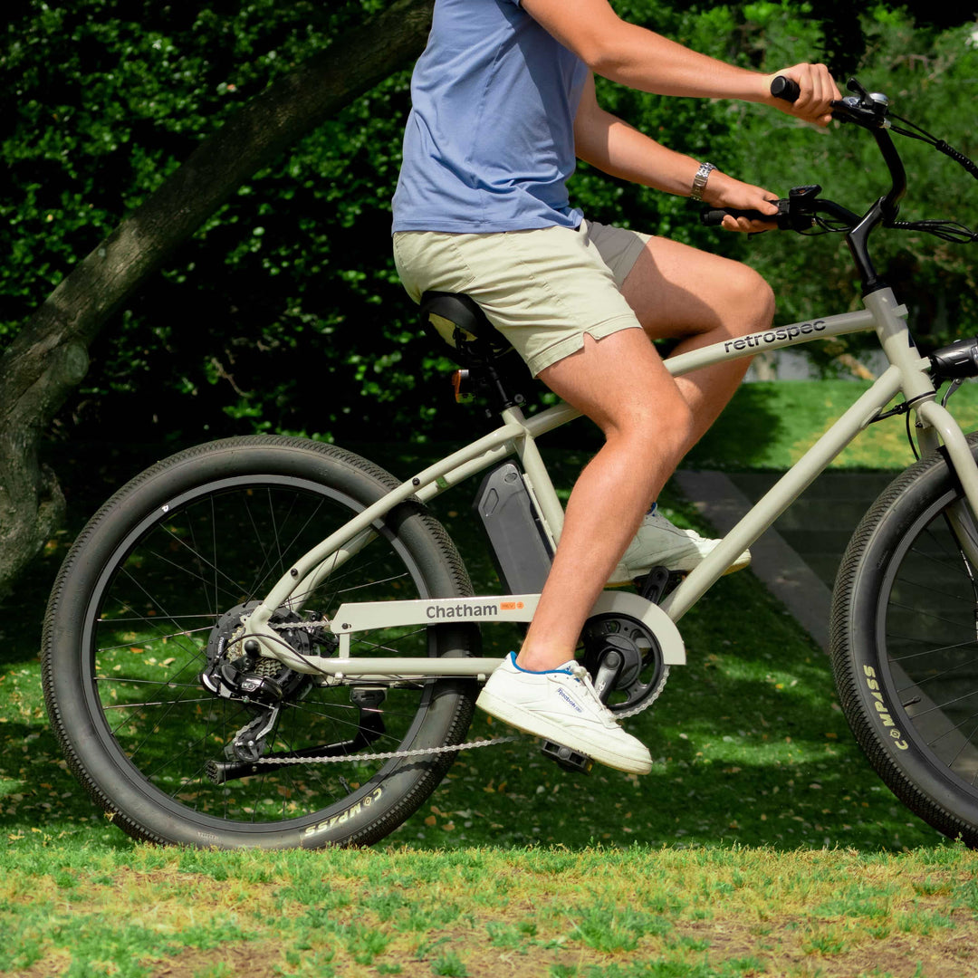Rider on a cream retrospec Chatham Rev electric cruiser bike, pedaling outdoors on grass with green foliage.