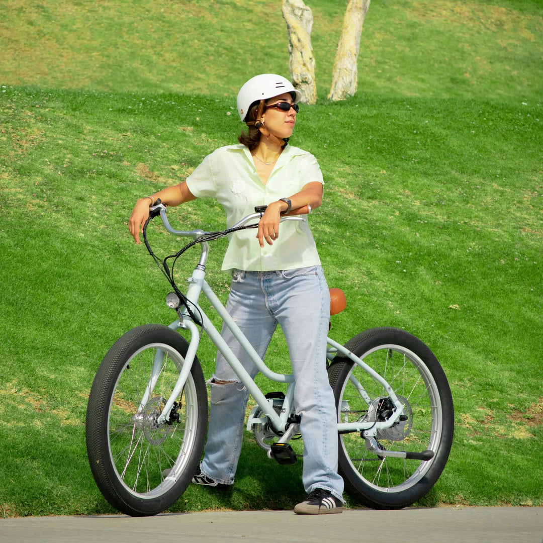 Woman in a white helmet leaning on a cream Retrospec electric cruiser bike on a sunny park path with a green lawn in the background.