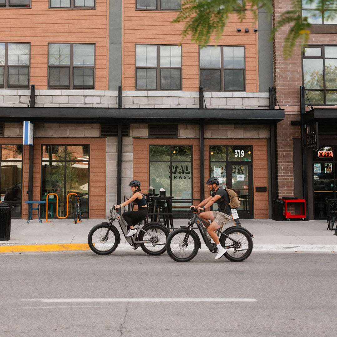 Two riders on light-colored retrospec electric bikes riding through downtown urban street storefront area.