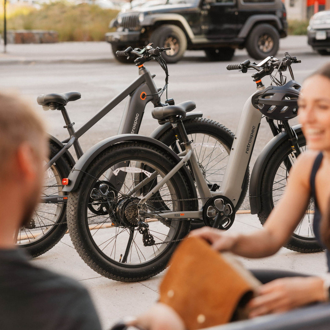 retrospec Koa Electric Bike in gray and black with orange accents, parked with multiple adult bikes in an urban outdoor setting.