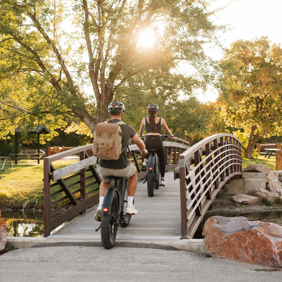 Two cyclists ride retrospec trail bikes across a wooden bridge over water surrounded by golden autumn foliage.