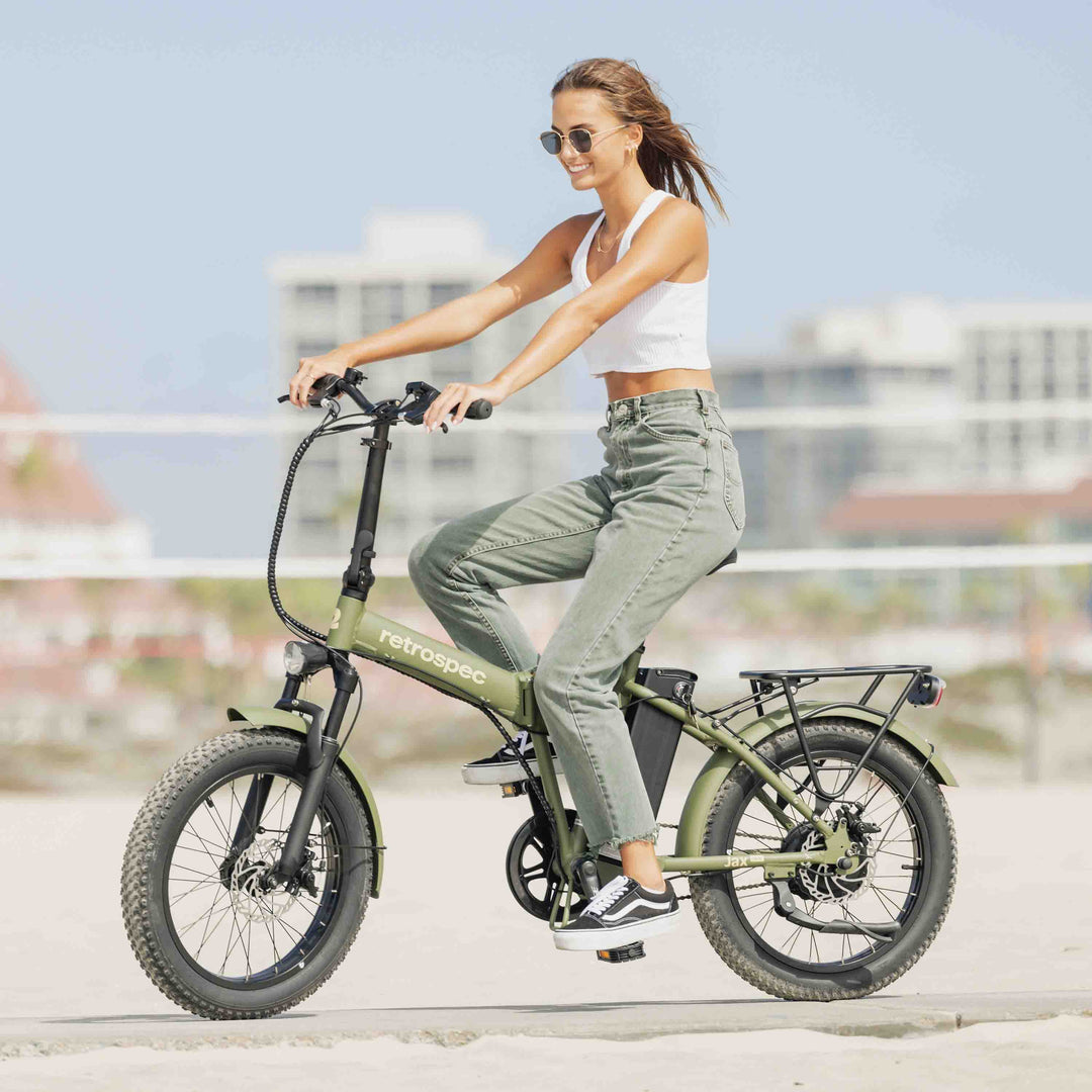 Woman riding sage green retrospec fat tire folding electric bike on urban rooftop with city skyline background.