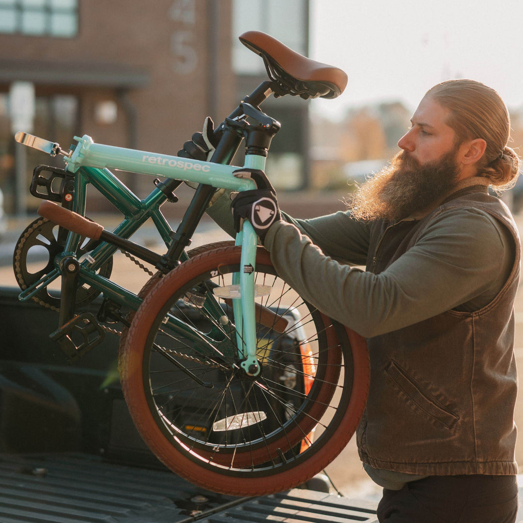 A person holds a folded retro-style bicycle with a mint green frame and brown tires, preparing to place it in a truck bed.