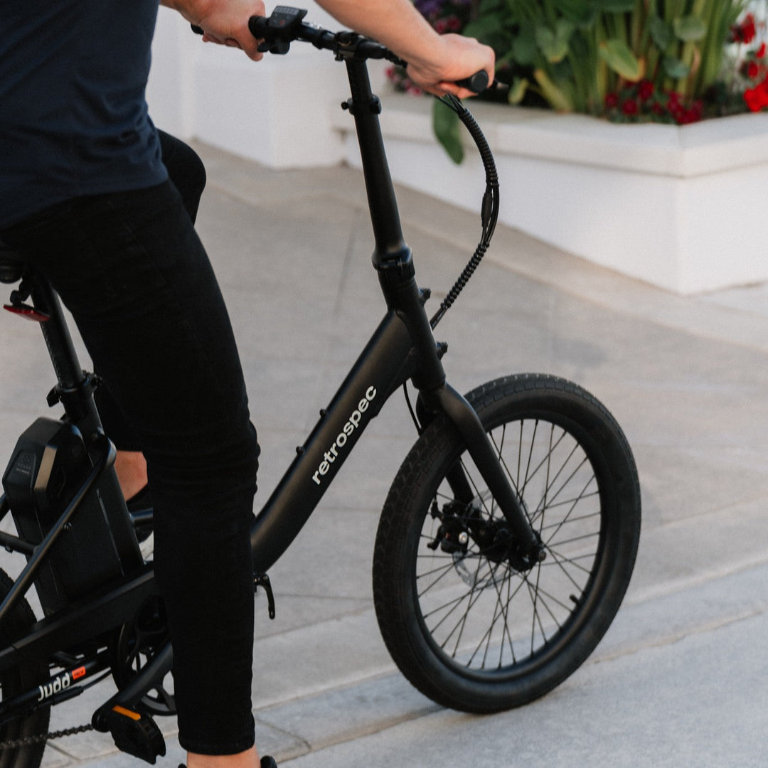 Close-up of a person riding a black Retrospec folding electric bike, gripping the handlebar, with a decorative flower bed in the background.