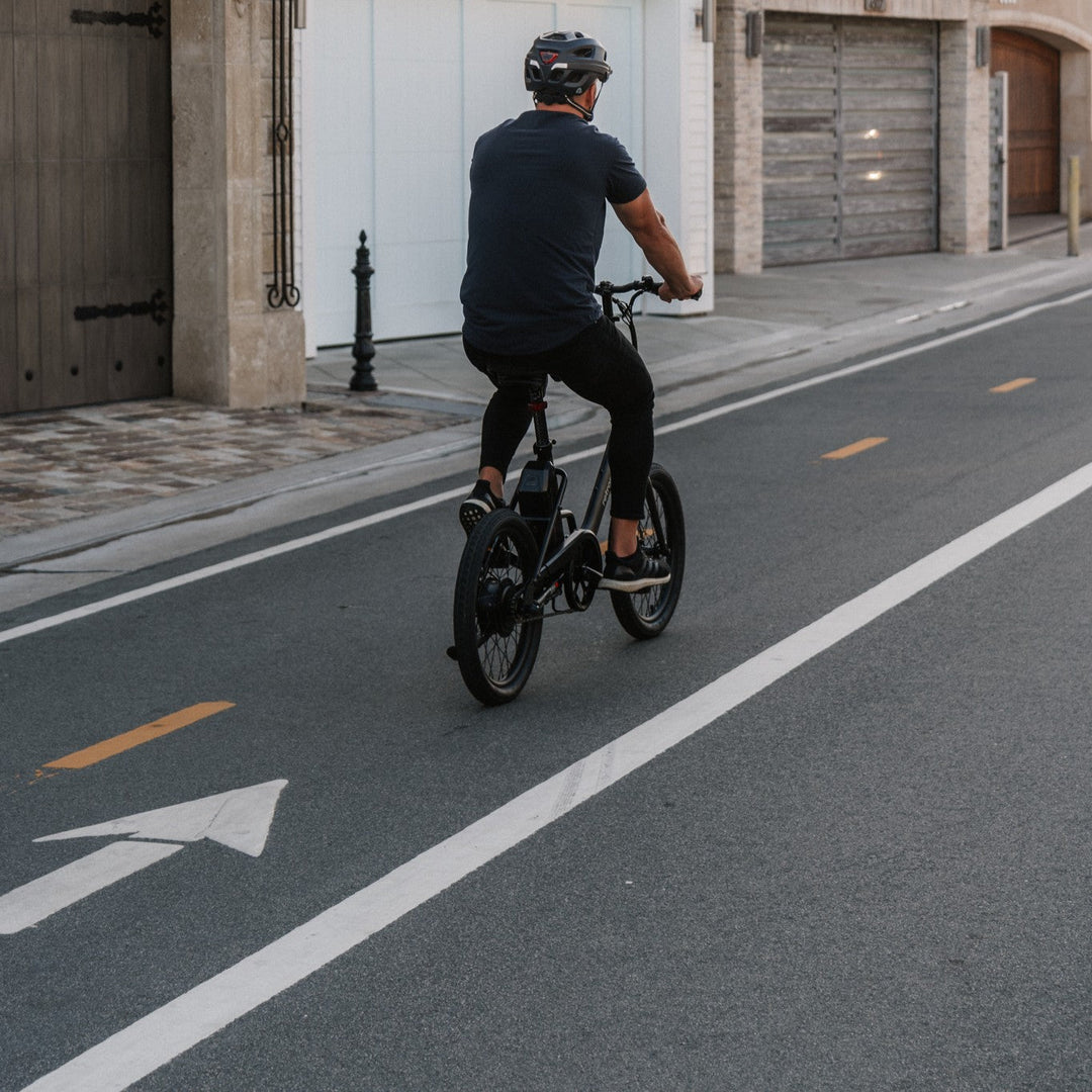 A cyclist in a helmet rides a black bike down a quiet street with arrow markings, passing residential garages and a lamp post.