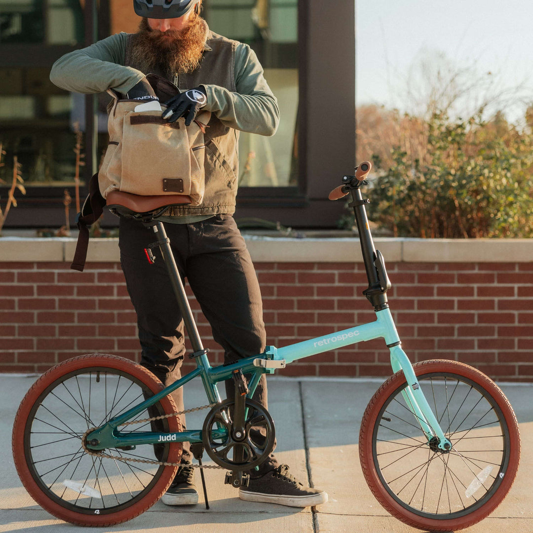 A bearded man in a helmet and gloves looks into his backpack while standing by a teal Judd folding bicycle on a sunny day.
