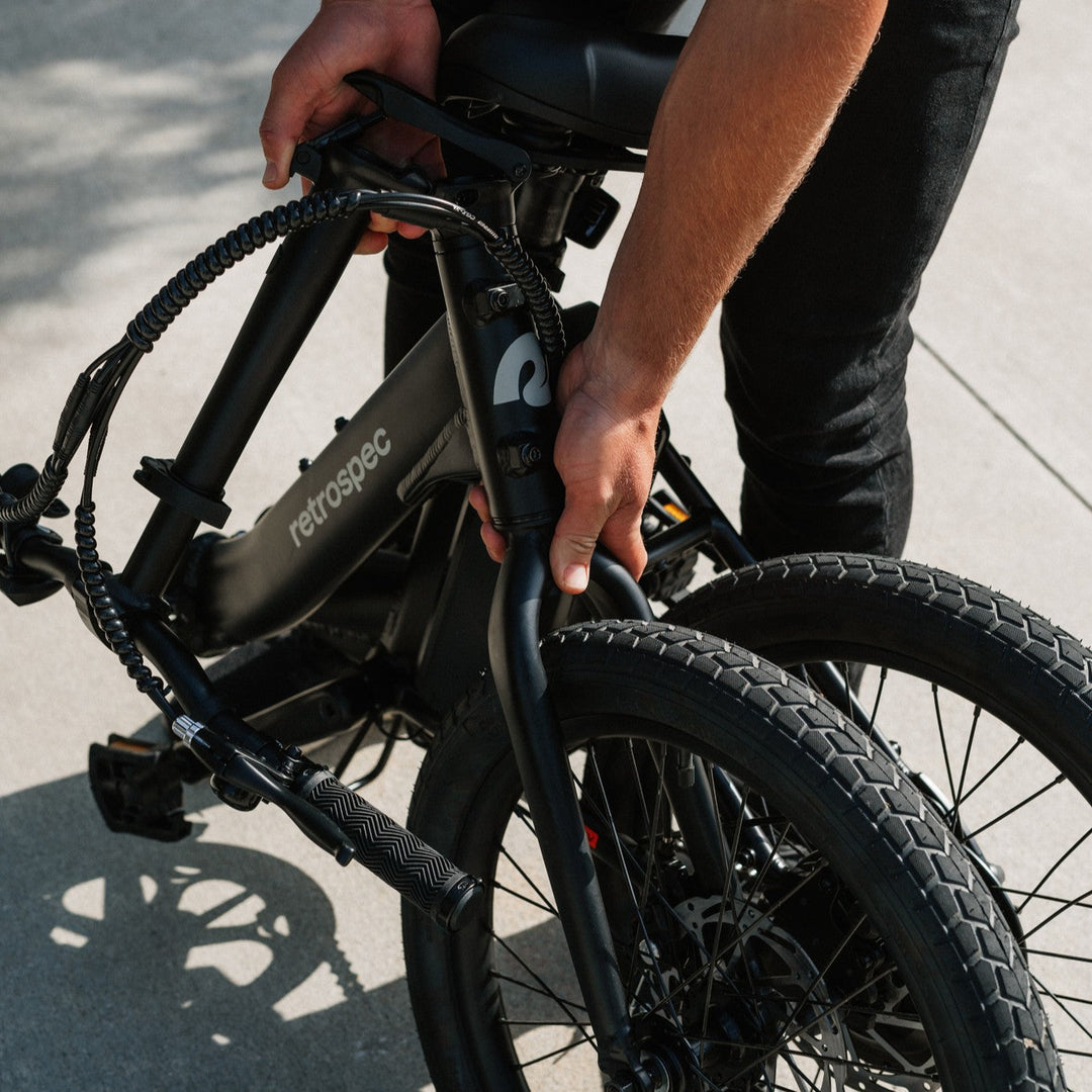 Rider holding retrospec Judd Rev folding ebike handlebars at beach with fat tires visible, lifestyle action shot