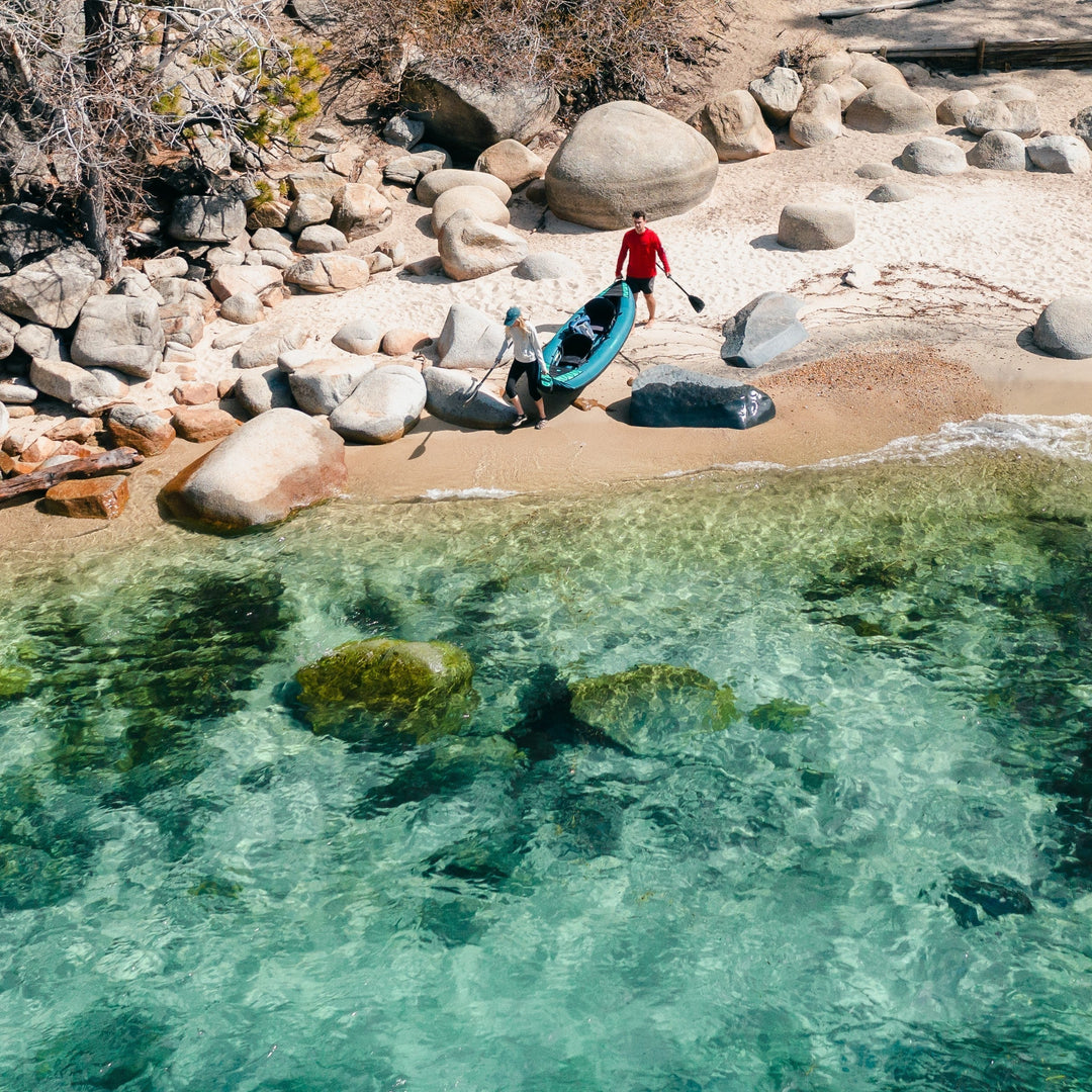 Two people on a sandy shore unload a kayak amid clear turquoise water and large rocks under a sunny sky.