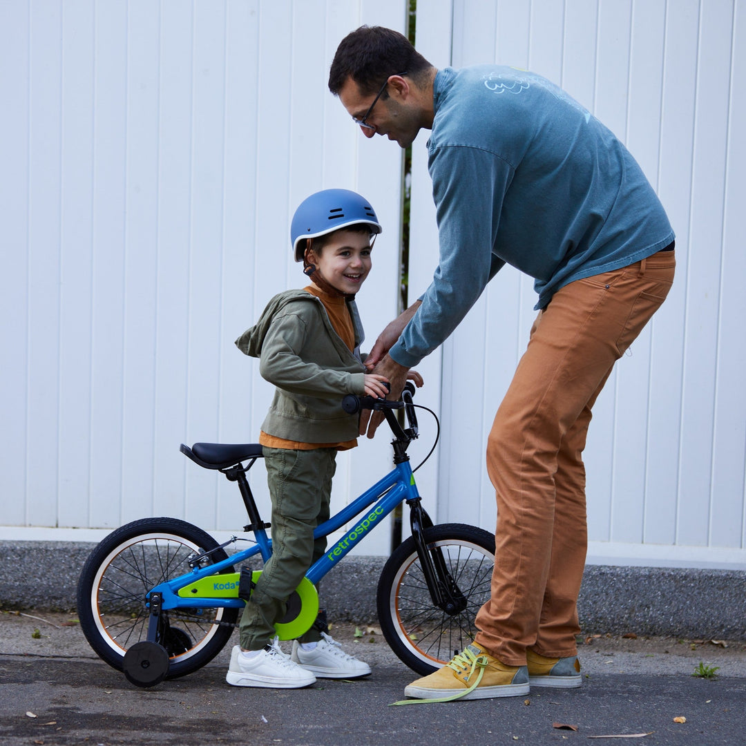 Adult teaching child to ride blue retrospec Koda kids pedal bike with training wheels and helmet outdoors.