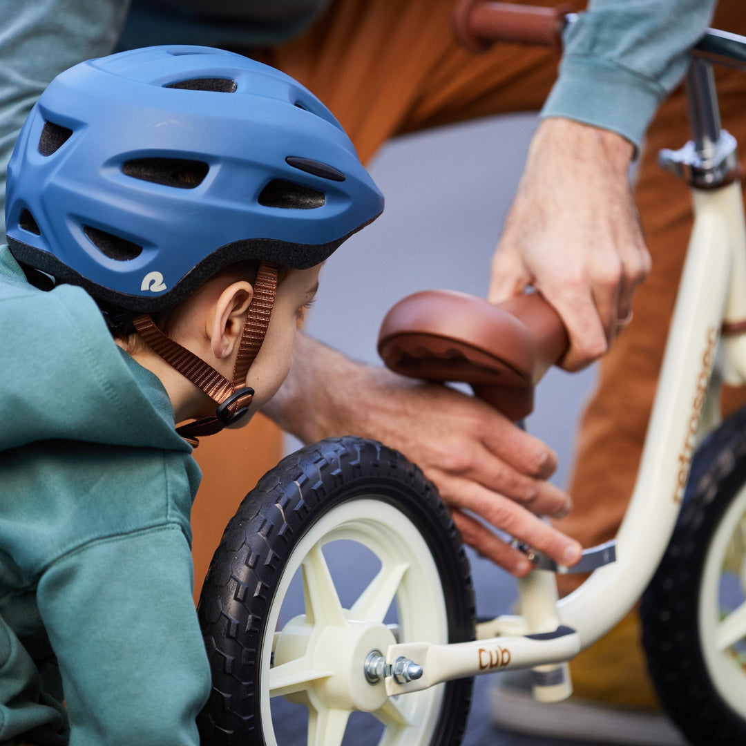 An adult adjusts the seat of a balance bike while a child wearing a blue helmet looks on.