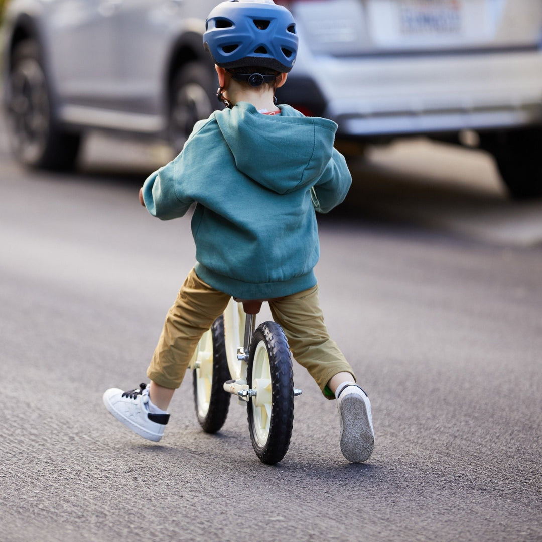A young child wearing a blue helmet rides a balance bike down a quiet street with a white car in the background.