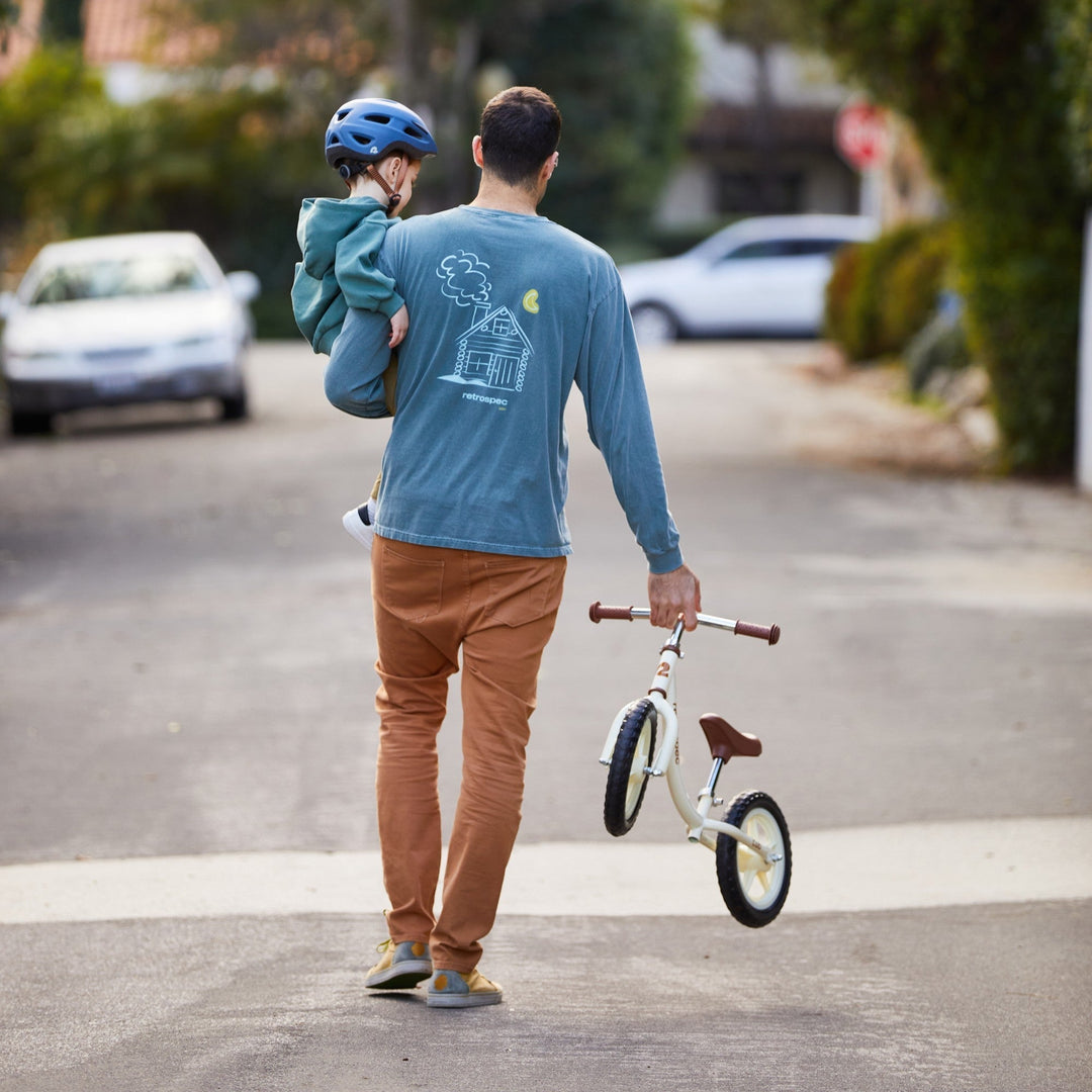 Parent carrying a young child on their shoulders while holding a white retrospec balance bike on a residential street