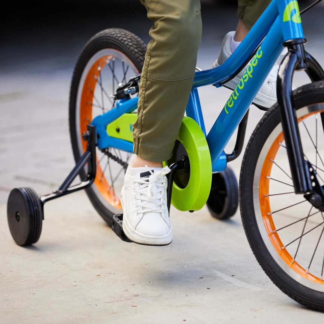 A child in olive green pants pedals a blue bike with orange wheels and a bright green chain guard, wearing white sneakers.