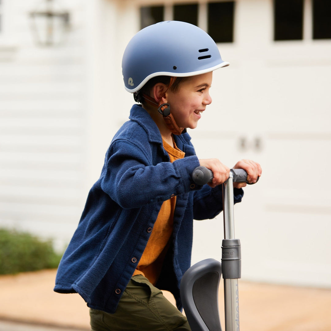 A child in a blue helmet rides a scooter, wearing a navy jacket over an orange shirt, outdoors in front of a white garage.