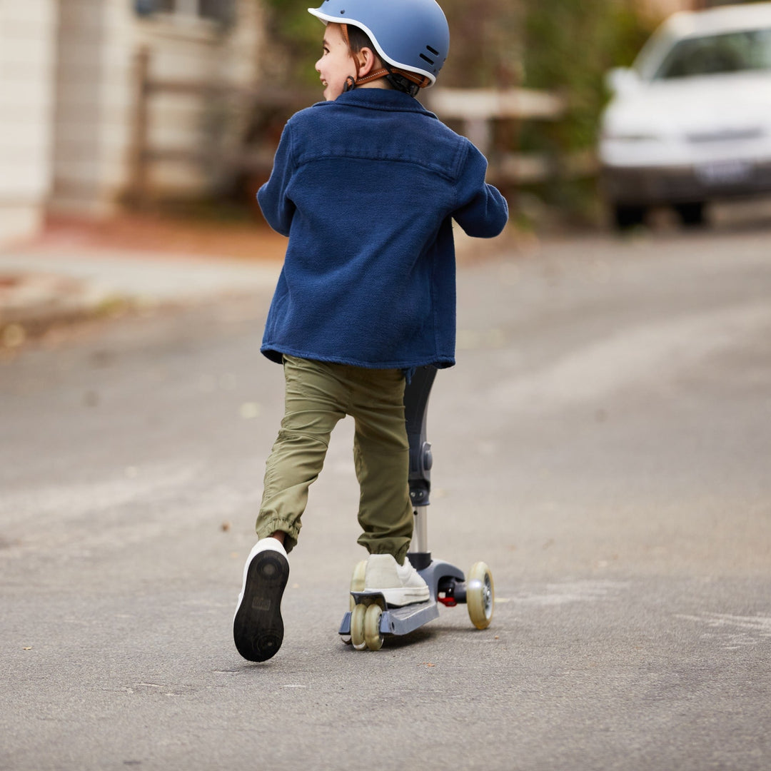 A child rides a scooter on a quiet street, wearing a blue jacket, green pants, and a helmet, enjoying a playful moment outdoors.