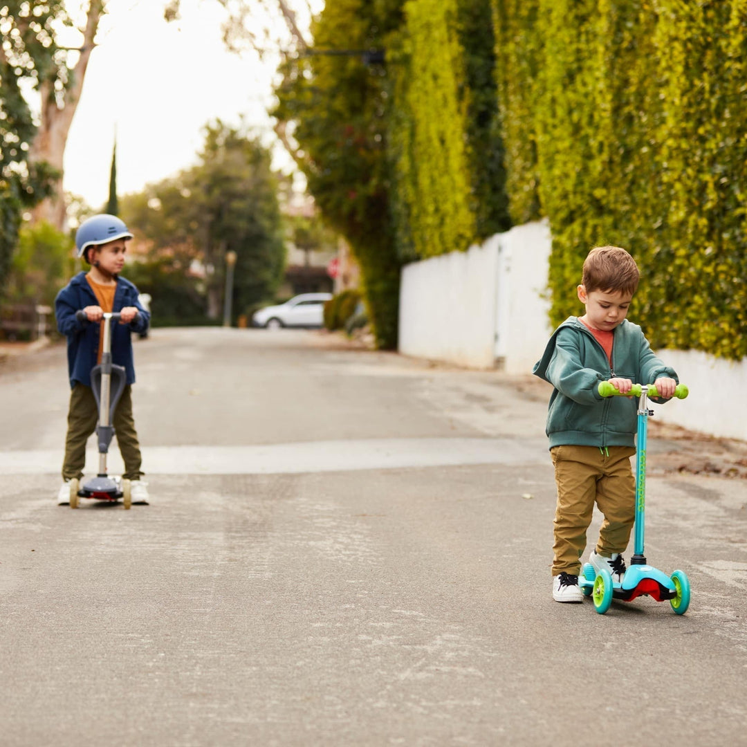 Two children riding scooters on a quiet street with tall green hedges.