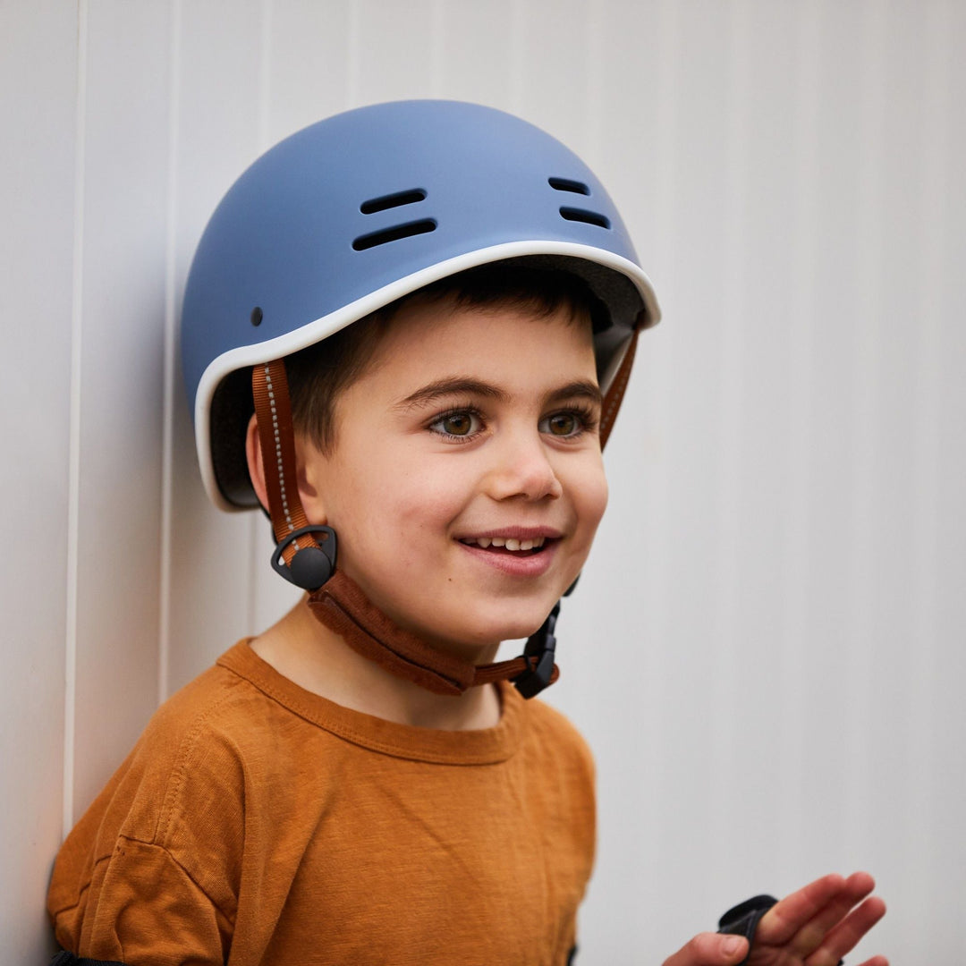 Child wearing a blue helmet and orange shirt leans against a white fence.