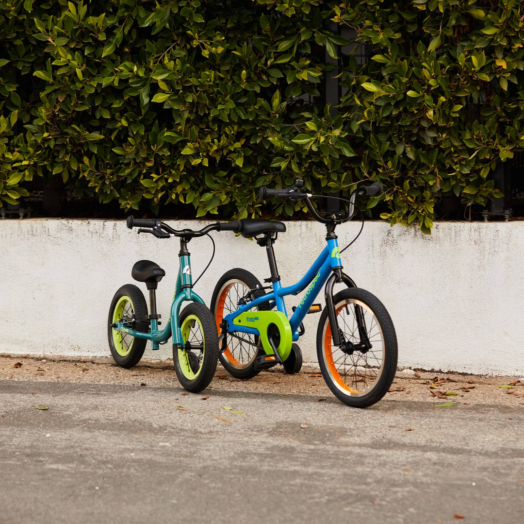 A balance bike and bike with training wheels standing beside one another in front of a white wall with green trees behind them.