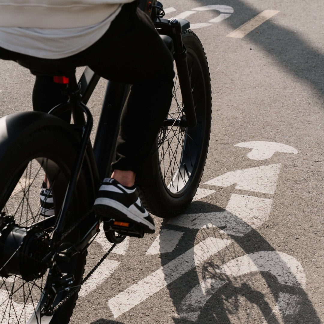 Low-angle view of a rider on a retrospec Koa Electric Bike in black, pedaling through a marked city bike lane with the fat rear tire and drivetrain visible in sharp detail.