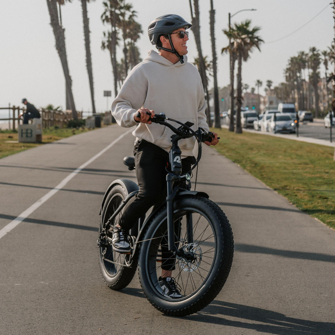 Man in sunglasses riding fat tire retrospec electric bike on coastal beach path lined with palm trees.