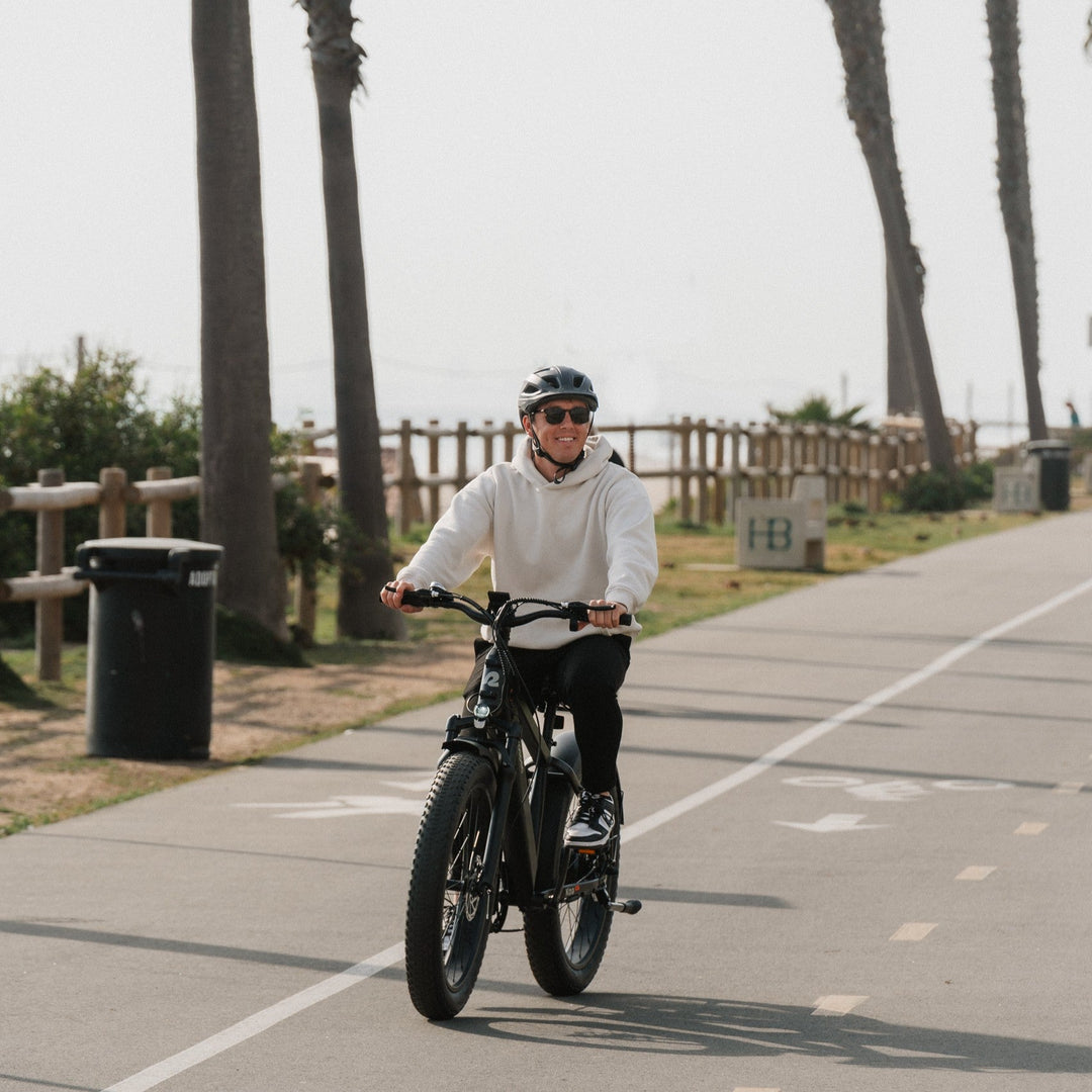 A person rides a black bicycle along a beachfront path with palm trees and a trash can visible in the background.