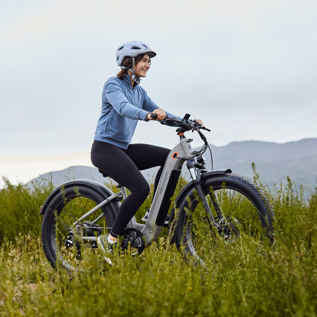 Woman in blue helmet riding a retrospec Koa Rev Plus all terrain electric bike through a green meadow with mountains