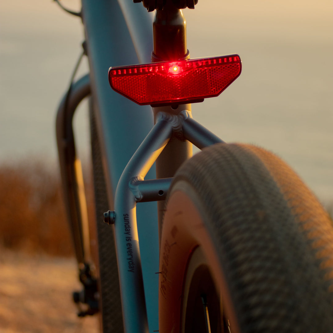 Close-up of the Retrospec Chatham Electric Bike's integrated red rear safety light glowing at golden hour, mounted on the seat stay above the brown-walled tire with the ocean visible in the background.