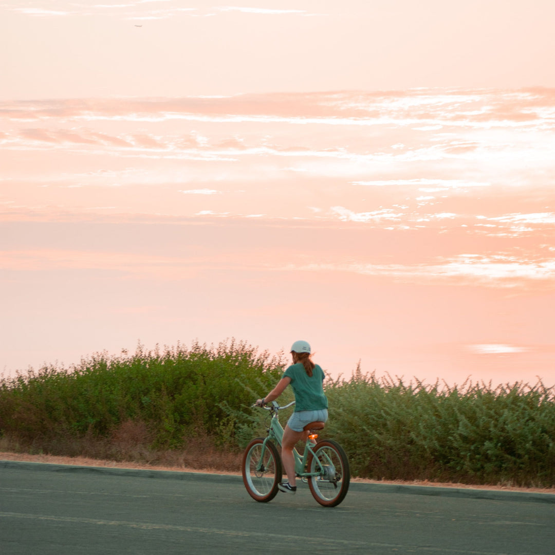 A woman rides a mint green electric beach cruiser bicycle along a road at sunset, with soft pink and orange skies and greenery in the background.