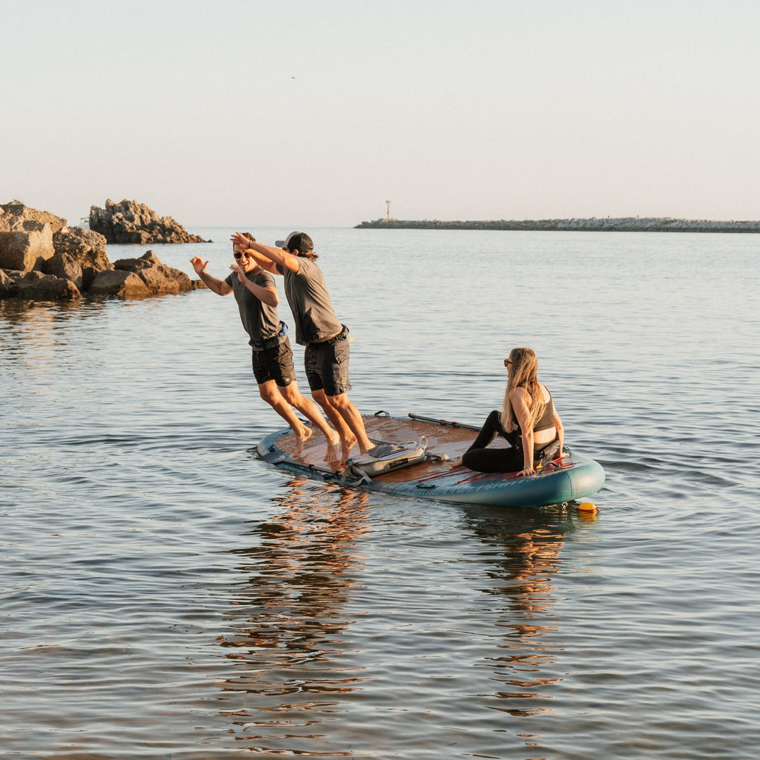 Two individuals are jumping off an inflatable paddleboard into calm water, while another sits on the board, enjoying the serene atmosphere.