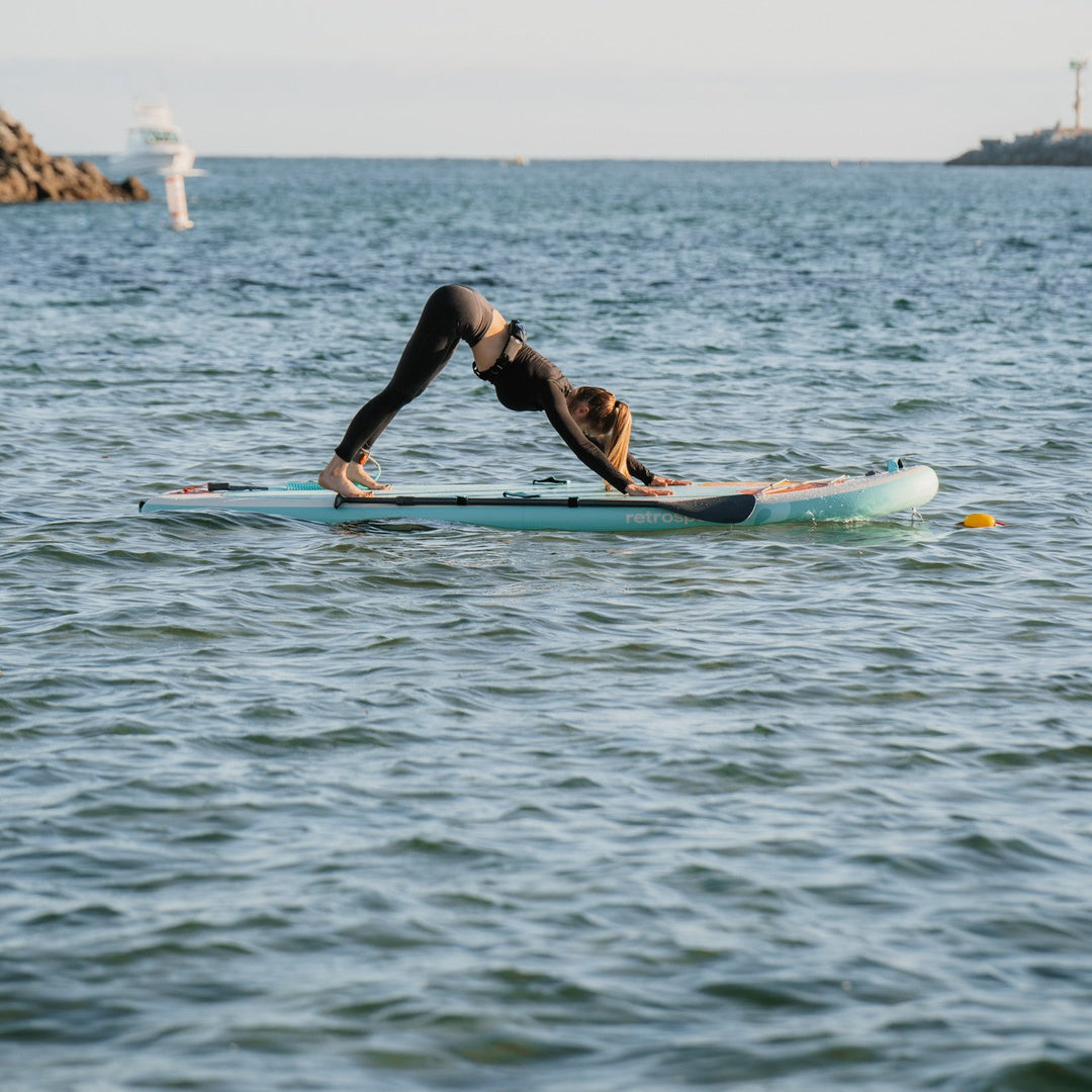 A person practicing yoga on a paddleboard, performing the downward dog pose on calm water with a coastal background.