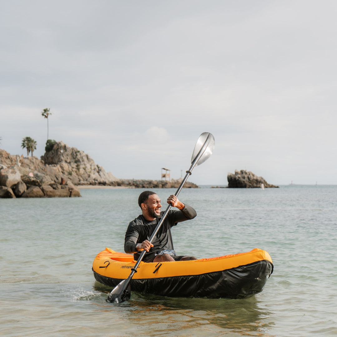 Man paddling retrospec Coaster one person kayak on turquoise water near rocky cliffs with palm trees.