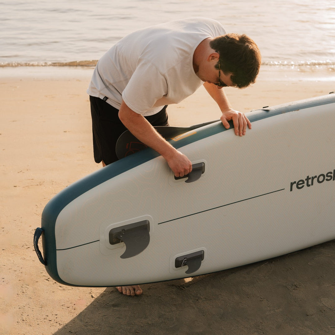 A person in a white t-shirt adjusts a paddleboard on the sandy beach, with calm waves in the background.